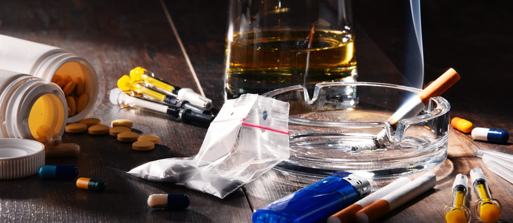 Various different pills, a white powder in a zip lock bag, an ash tray with a cigarette butt, a glass of dark liquor, and two syringes. 